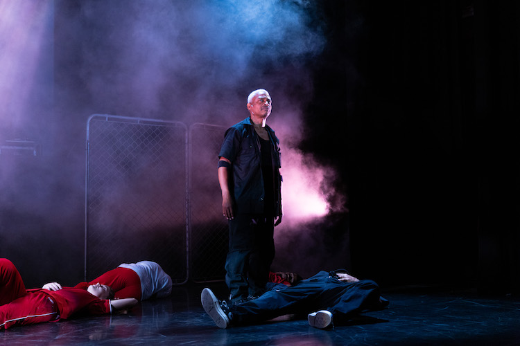 a Black man with blonde short hair in center of a smoke filled stage, bodies tragically lying prone around him.